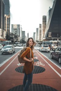 Young white woman smiling while standing in a crosswalk on a busy city street. She has dark blonde hair and is wearing a dark orange tunic, blue jeans, and a tan backpack.