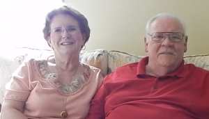 Image shows an older white couple smiling and sitting side by side on a loveseat. The woman has dark red hair and wears glasses. She's wearing a peach shirt with floral stitching around the collar and a long, gold chain with a round locket. The man has a receding hairline with white hair and mustache. He wears glasses and a red, collared shirt.